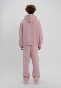 Personne debout, de dos, portant un sweat à capuche rose ample et un pantalon assorti avec des baskets blanches sur un fond blanc.