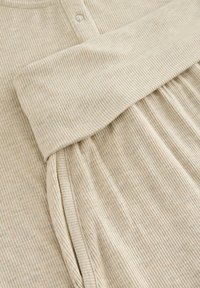 Tissu beige à nervures avec une surface texturée, présentant un col arrondi et une fermeture à bouton. Design doux et fluide avec des détails froncés.
