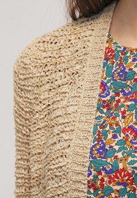 Beige stickad cardigan med ett texturerat mönster, bärs över en färgglad blus med blommönster som har olika nyanser av blått, rött och grönt.