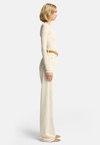 Beige Langarmoberteil, kombiniert mit hochgeschnittenen, locker sitzenden cremefarbenen Hosen und einem braunen Gürtel. Braune Schuhe mit Nieten runden das Outfit ab.