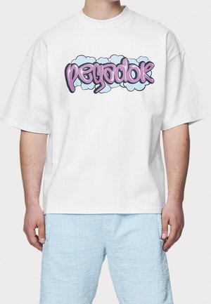 Hombre vestido con camiseta blanca de gran tamaño con texto estilo grafiti en púrpura y pantalones cortos texturizados azul claro, de pie frente a un fondo liso.