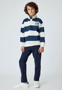 Marinblå och vit randig tröja med hög krage och dragkedja, med grön text. Tillsammans med marinblå sweatpants och vita sneakers.