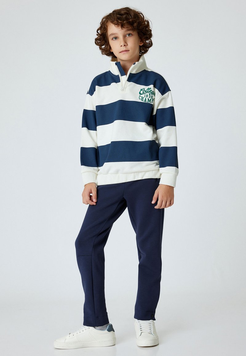 Marinblå och vit randig tröja med hög krage och dragkedja, med grön text. Tillsammans med marinblå sweatpants och vita sneakers.
