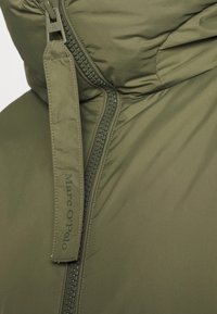 Olivgrön puffjacka med dragkedja. Har en märkesdragkedja med "Marc O'Polo" och texturerad dragkedja. Slät, isolerad material.