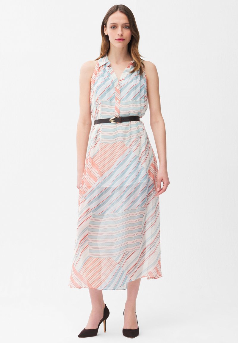 Frau trägt ein ärmelloses, gestreiftes Midikleid in Pastellfarben, das mit einem schwarzen Gürtel tailliert ist, kombiniert mit schwarzen High Heels, steht vor einem weißen Hintergrund.