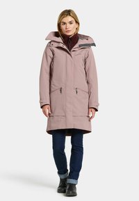 Abrigo impermeable de color rosa claro con un cuello alto, bolsillos con cremallera y capucha ajustable. Combinado con pantalones vaqueros oscuros y botas de tobillo negras.