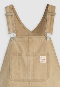 Salopette en toile beige avec clips en métal réglables, dotée d'une grande poche avant et d'un patch logo sur le côté. Texture lisse.