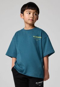 Turkis oversized bomulds t-shirt med korte ærmer og frontlogo i gul. Parret med sorte bukser med hvid branding ved kanten.