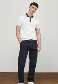 Young man standing with hands in pockets wearing white short-sleeve polo shirt and dark blue trousers on wooden floor.