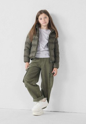 Olijfgroene pufferjack, grijs T-shirt, olijfgroene cargobroek met zakken en witte platform sneakers. Staande tegen een lichte achtergrond.
