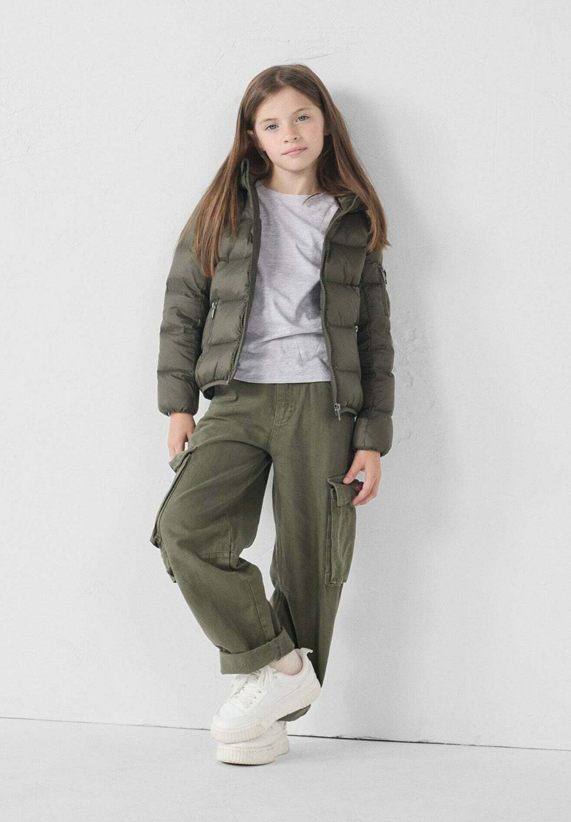 Olijfgroene pufferjack, grijs T-shirt, olijfgroene cargobroek met zakken en witte platform sneakers. Staande tegen een lichte achtergrond.
