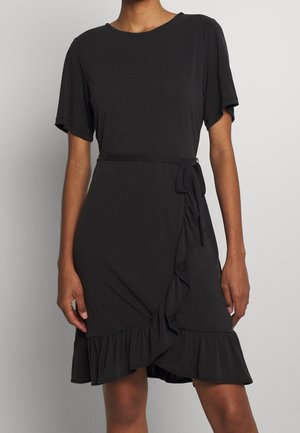 Femme portant une robe portefeuille noire à manches courtes, avec un ourlet à volants et un lien latéral à la taille.