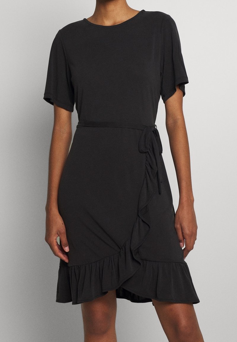 Femme portant une robe portefeuille noire à manches courtes, avec un ourlet à volants et un lien latéral à la taille.