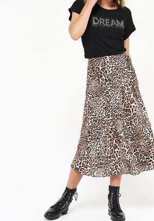 Mujer con camiseta negra con la palabra "DREAM", falda midi de estampado de leopardo y botas negras con cordones, de pie sobre un fondo blanco.