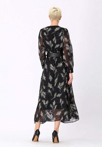 Personne avec des cheveux blonds courts portant une robe noire transparente avec des motifs de plumes blanches, une ceinture noire et des talons hauts noirs, se tenant de dos.