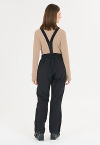 Kvinde iført beige langærmet top, sorte skiløb bukser med seler og sorte skistøvler, står vendt væk mod en hvid baggrund.