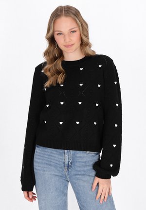 Maglione nero lavorato a maglia con un motivo a diamante e piccoli dettagli a forma di cuore bianchi. Maniche lunghe con polsini a coste. Abbinato a jeans azzurri chiari.
