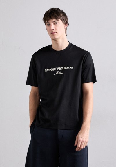 Camiseta de algodón negra con un logo central de "Emporio Armani" en blanco y "Milano" debajo, de mangas cortas y cuello redondo.