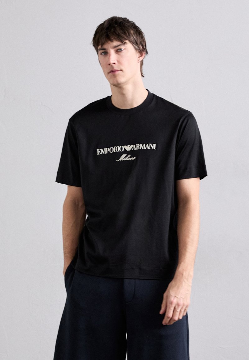 T-shirt en coton noir avec un logo central "Emporio Armani" en blanc et "Milano" en dessous, manches courtes et col rond.