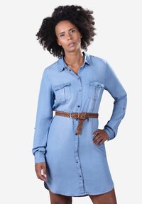 Mujer con cabello rizado que lleva un vestido de mezclilla azul claro de manga larga con bolsillos en el pecho y un cinturón marrón tejido, posando con la mano en la cadera.