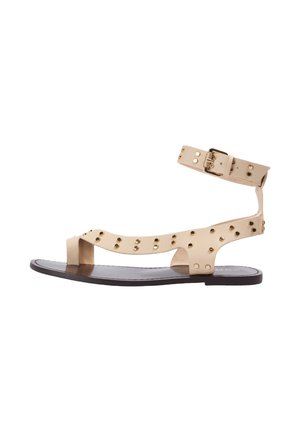 Sandalia plana beige con tira en el tobillo, hebilla dorada y tachuelas doradas en las correas, y una suela marrón oscuro.