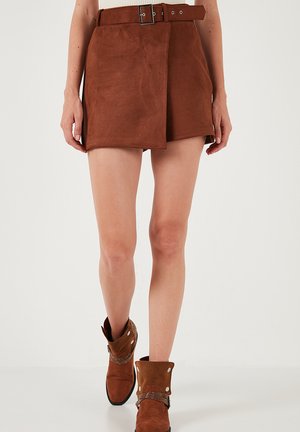Jupe-culotte en daim marron avec ceinture, présentant un design enveloppant. Associée à des bottines marron, mettant en valeur des accessoires décoratifs et un détail replié.