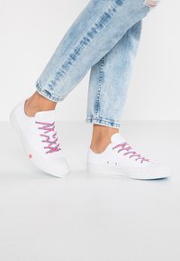 Białe trampki z różowymi i niebieskimi sznurówkami, gumową podeszwą, okrągłym czubkiem oraz różową gwiazdką na boku. Noszone z jasnoniebieskimi dżinsami.