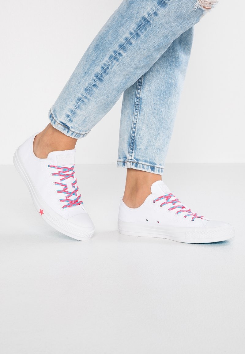 Białe trampki z różowymi i niebieskimi sznurówkami, gumową podeszwą, okrągłym czubkiem oraz różową gwiazdką na boku. Noszone z jasnoniebieskimi dżinsami.