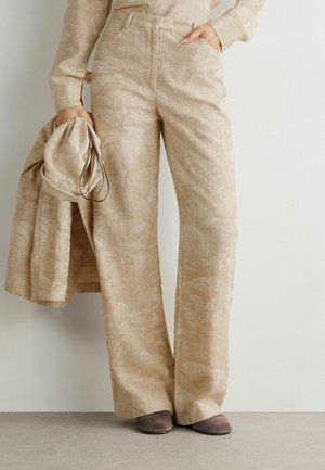 Frau trägt beige gemusterte, hochtaillierte, weit geschnittene Hosen und passende Jacke, hält einen Hut und steht vor einer weißen Wand.