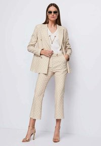 Completo beige con motivo, giacca doppiopetto e pantaloni cropped su misura. Indossato sopra una maglietta bianca, abbinato a accessori dorati e tacchi.