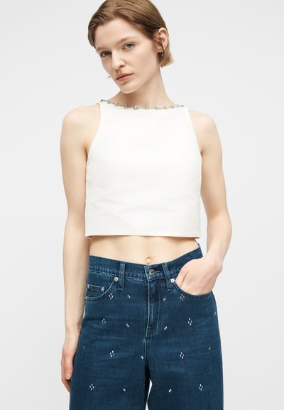 Femme portant un haut court sans manches blanc avec un col orné de perles et un jean taille haute bleu avec de petites embellissements argentées, main dans la poche.