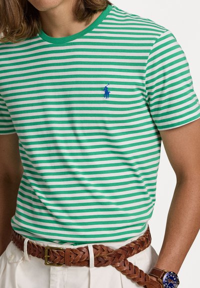 Πράσινο και λευκό ριγέ μπλουζάκι με στρογγυλή λαιμόκοψη και κεντημένο μπλε λογότυπο. Συνδυασμένο με καφέ πλέκτης ζώνη πάνω από λευκά σορτς.