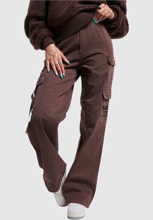 Pantalones cargo marrones de pierna ancha, con múltiples bolsillos, un tejido texturizado y detalles gráficos impresos. Combinados con zapatillas blancas.