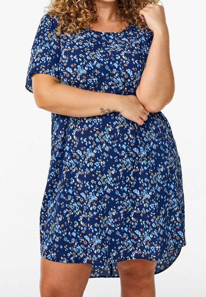 Femme aux cheveux bouclés portant une robe bleu marine à manches courtes avec un petit imprimé floral, les bras croisés.