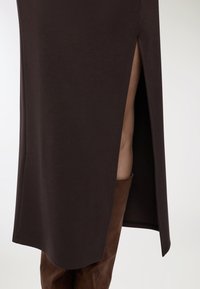 Campo ravvicinato di una gonna marrone scuro con uno spacco laterale alto, che rivela una gamba che indossa un stivale in pelle marrone fino al ginocchio.