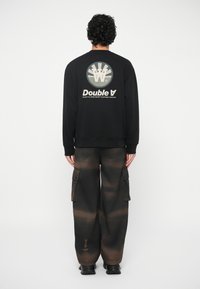 Μαύρη μπλούζα με κουκούλα με κυκλικό γραφικό και το κείμενο "Double A" στο πίσω μέρος, συνδυασμένη με σκούρες cargo παντελόνες που διαθέτουν gradient σχέδιο.