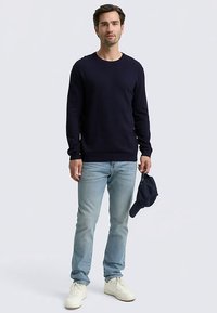 Granatowy sweter z dzianiny z długimi rękawami i okrągłym dekoltem; noszony z jasnoniebieskimi dżinsami i białymi trampkami; trzyma ciemną czapkę.