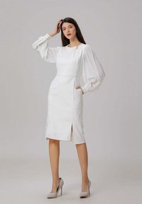 Robe blanche ajustée avec des manches ballon, fentes latérales et poches. Texture lisse et design épuré, associée à des escarpins beiges clairs.