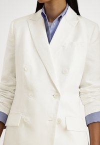 Λευκό σακάκι διπλής σειράς κουμπιών από λινό, με δύο κουμπιά, πέτα με εγκοπές και μανίκια γυρισμένα, φορεμένο πάνω από ανοιχτό μπλε πουκάμισο.