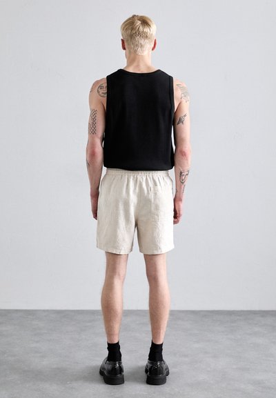 Haut sans manches noir, shorts en lin beige avec taille élastique et chaussures noires. Tatouages visibles sur les bras. Arrière-plan neutre.