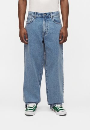 Άνδρας που φορά ανοιχτό μπλε τζιν με φαρδιά μπατζάκια, λευκό πουκάμισο και πράσινα αθλητικά παπούτσια, στέκεται μπροστά σε απλό λευκό φόντο.