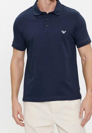 Mand iført en marineblå poloshirt med korte ærmer, tre-knaps slids og lille hvidt logo på venstre bryst, kombineret med lyse beige bukser.