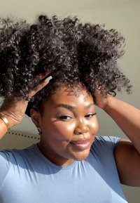 Capelli neri ricci sollevati con le mani; indossa una camicia azzurra chiara. Pelle liscia con un leggero luccichio, illuminazione intensa che evidenzia i tratti.