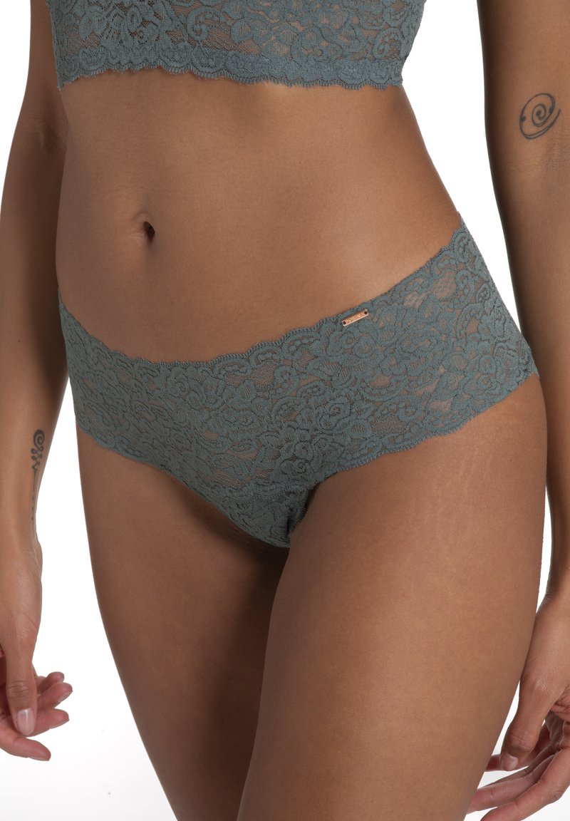 Tirkīza mežģīņu hipster apakšveļa ar viļņotām malām, kurai ir ziedu raksts un neliels metalizēts logotipa akcents. Mīksta, caurspīdīga tekstūra.