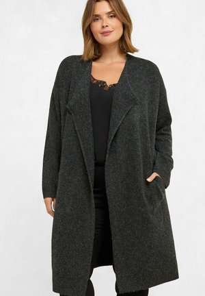 Frau trägt eine lange dunkelgraue offene Strickjacke über einem schwarzen Top mit Spitzenbesatz und steht mit einer Hand in der Tasche.