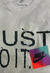 T-shirt bianco in cotone con la scritta audace e nera "JUST DO IT" in stile rovinato. Include un'etichetta Nike quadrata multicolore nell'angolo in basso.