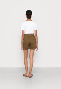 T-shirt blanche à manches courtes associée à un short à taille élastique vert olive. Le modèle porte des sandales marron et se tient sur un sol blanc.