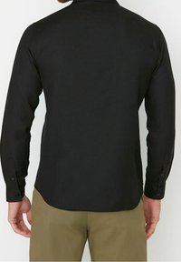 Zwarte langesleeve shirt van gladde stof, met een afgeronde zoom en met knopen afgesloten manchetten. De achterkant toont een eenvoudig, klassiek ontwerp.