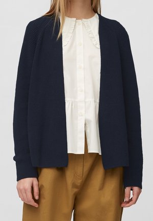 Kobieta w granatowym dzianinowym kardiganie noszonym na białej bluzce z falbankowym kołnierzem zapinanej na guziki oraz musztardowo-brązowych spodniach z wysokim stanem.