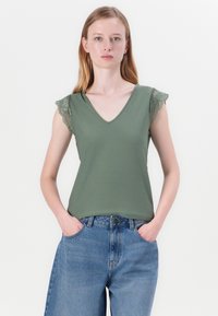 Grønn V-hals topp med blonder på korte ermer, kombinert med høyhalsa blå denimshorts. Stoffet virker glatt og tettsittende.